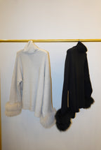 Two folded sweaters, one beige and one black with a fur pom-pom, on a white background.